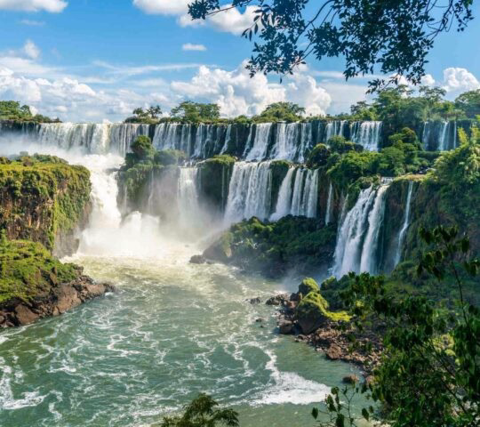 A wide, powerful waterfall with multiple tiers of white water cascading down lush green cliffs under a blue sky.