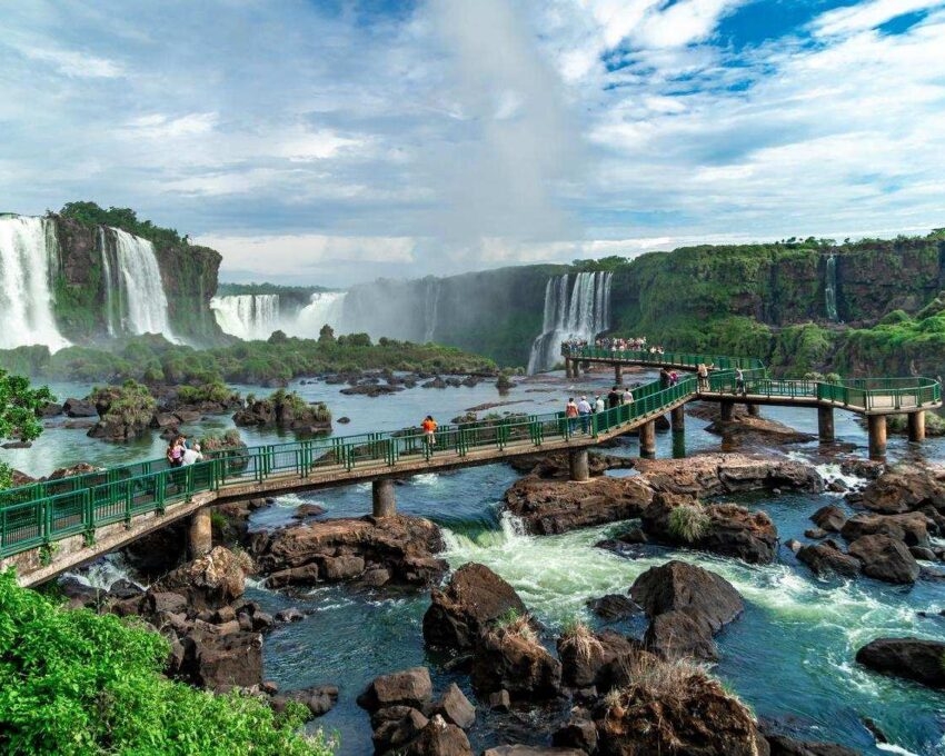 A scenic view of Iguazu Falls with white water cascading over cliffs and a pedestrian walkway across the river.