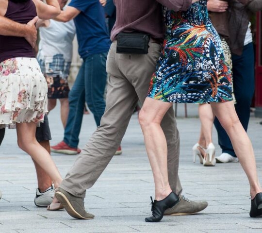 Close-up of several couples dancing tango on a stone-paved public square, focusing on their legs and footwear.