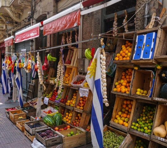 An outdoor fruit and vegetable market stall with wooden crates of produce and Uruguayan flags hanging.