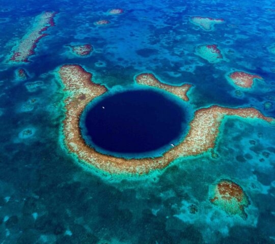 Aerial view of a large circular deep blue marine sinkhole surrounded by a shallow turquoise coral reef.