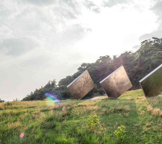 Three large square metallic sculptures on a green hill reflecting the sky and surroundings.