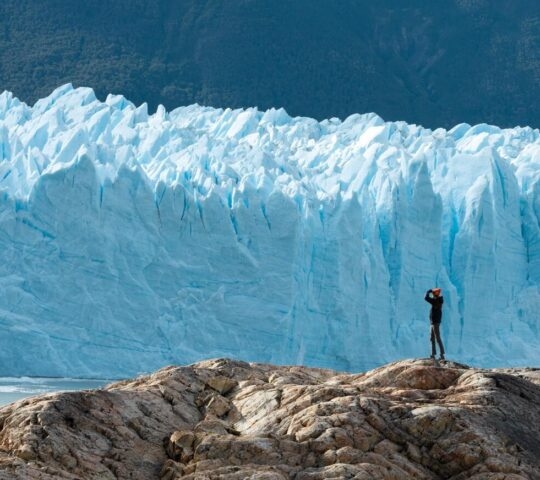 Small figure of a person standing on rocks in front of a giant, textured blue ice wall of a glacier.