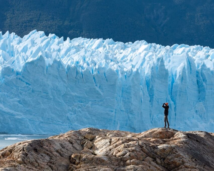 Small figure of a person standing on rocks in front of a giant, textured blue ice wall of a glacier.