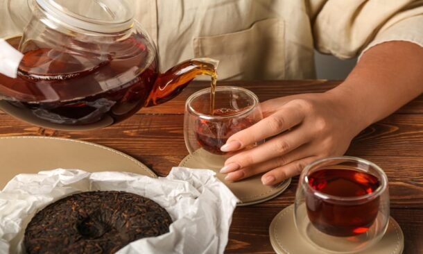 Dark tea being poured from a glass teapot into a small glass cup on a wooden table.