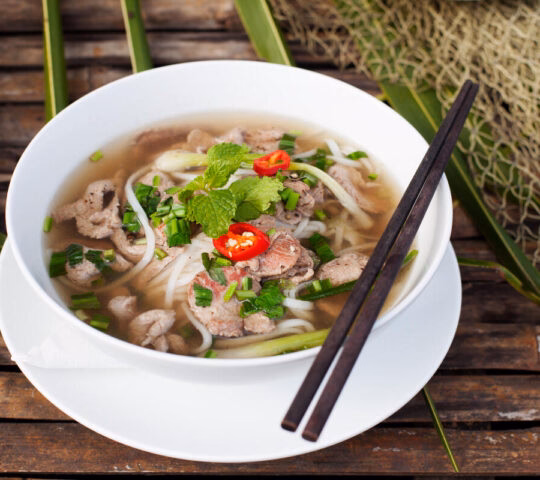 A bowl of steaming noodle soup with sliced meat, greens, and chilies with chopsticks.