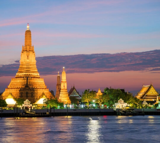 A large, ornate Buddhist temple lit up at dusk next to a river.