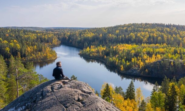 A man sits on a large rock looking out over a blue lake and yellow autumn trees.