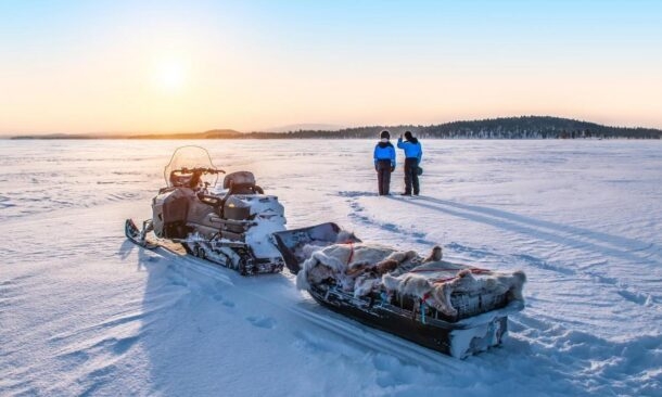 A snowmobile and sled on a vast snowy plain with two figures standing in the distance at dawn.