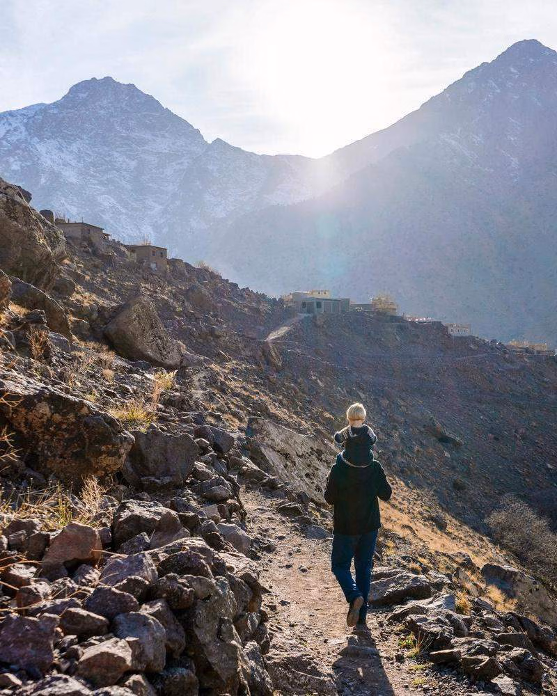 A hiker with a child on their shoulders walks along a steep, rocky path with snow-capped mountains in the background.