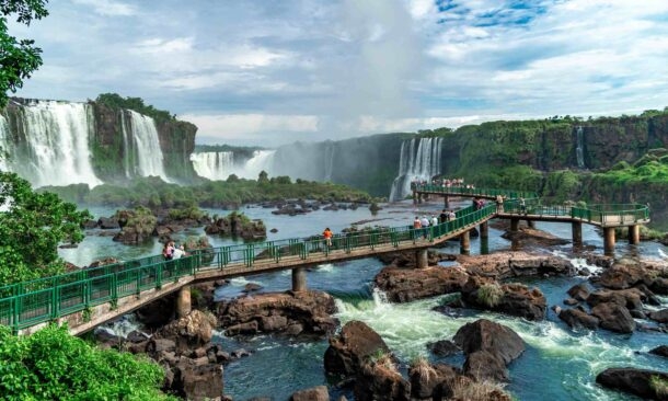 A system of walkways over a river leading toward several large, powerful waterfalls in a forest.