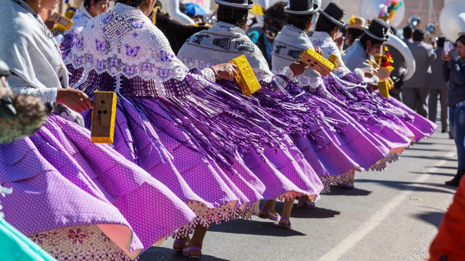 Dancers in purple skirts and white lace shawls perform in a parade, holding small wooden instruments.