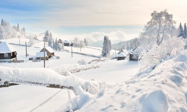 A snowy winter village landscape with scattered wooden houses, covered trees, and hills under a bright sky.
