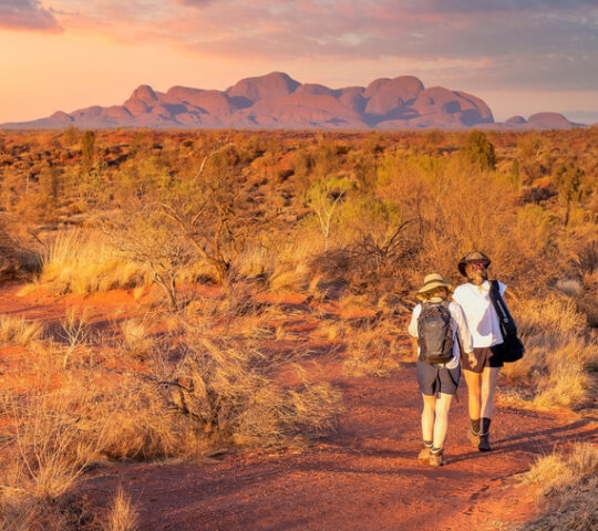 Two people walk on a red trail toward large rounded rock formations under a soft pink and blue sunset sky.