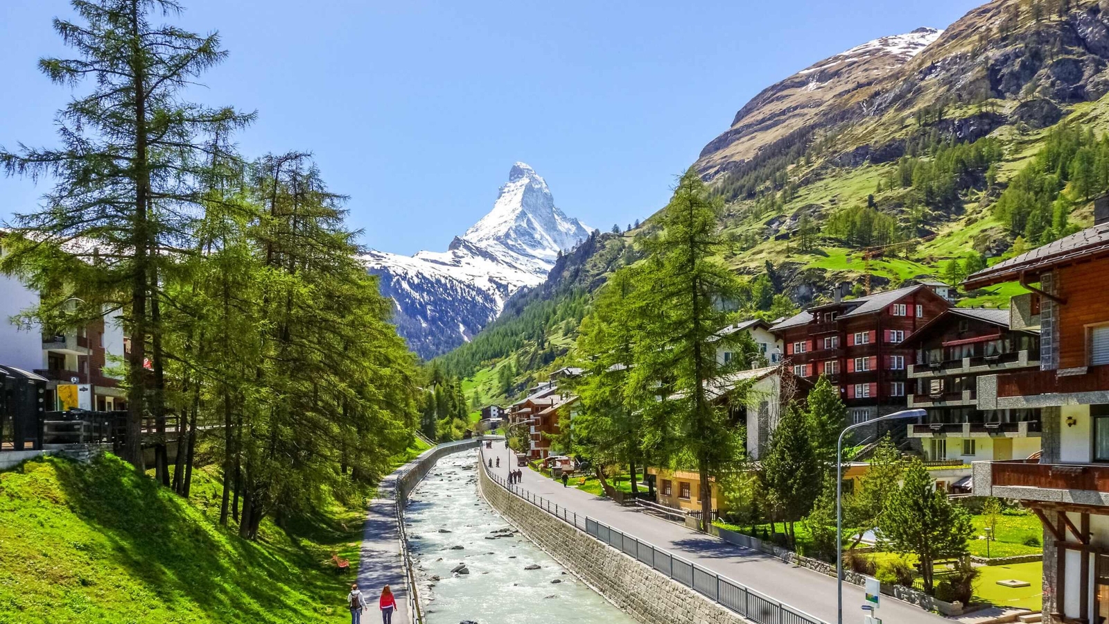 An alpine town with a stream and pine trees, featuring a prominent snow-capped mountain peak in the distance.