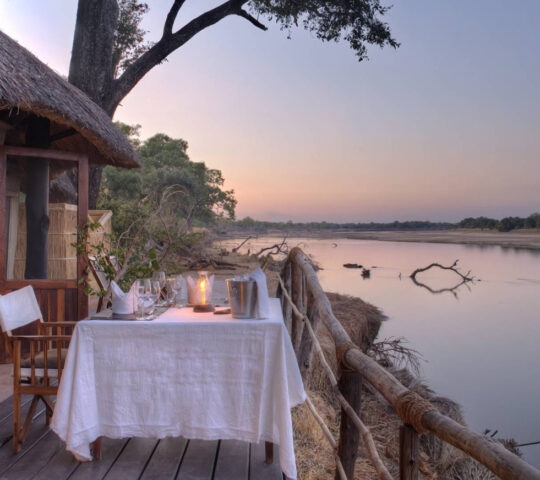 Exterior image of outdoor dining area, with small table and two chairs, set with a white table cloth, ice bucket, glasses and a candle. The view looks over a still river