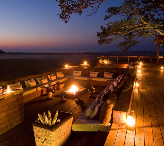 Exterior image of outdoor seating area on a wooden verandah. Comfy chairs around fire pits with softly glowing lanterns