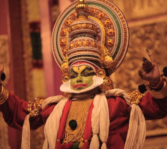 A dancer in traditional clothing, mask, and headdress dances on stage