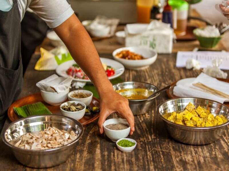 Close-up of a person's hand reaching for a small white bowl among several metal bowls filled with food on a wooden table.