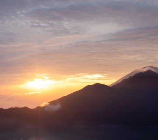 A golden sunrise behind dark mountain silhouettes with soft clouds in the morning sky.