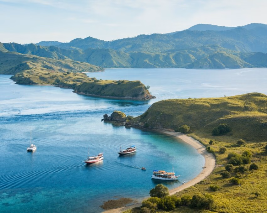 Aerial view of several boats anchored in a calm turquoise bay surrounded by rolling green hills under a bright sky.