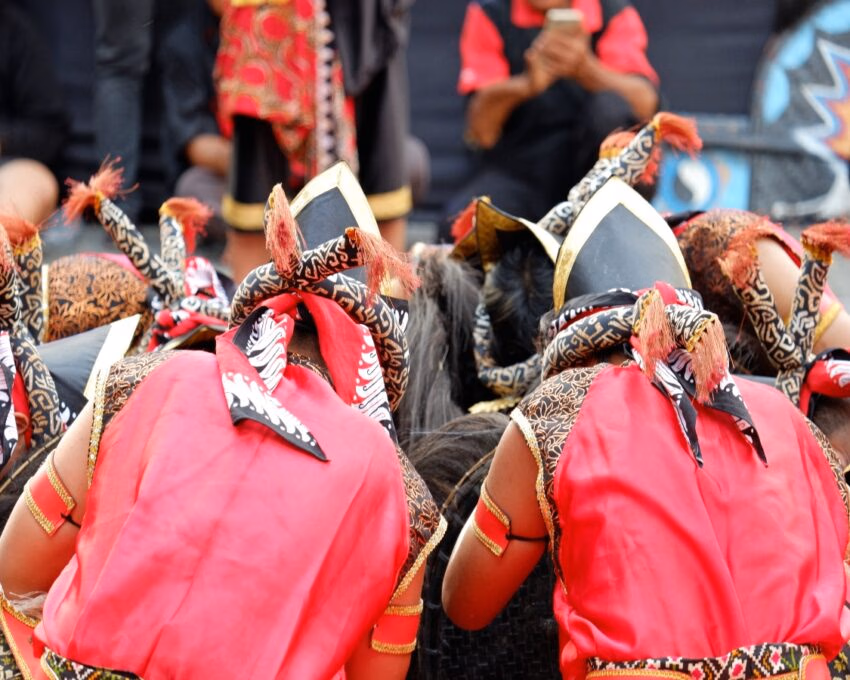 Back view of several people in red traditional clothing and headpieces bowing forward in a group.