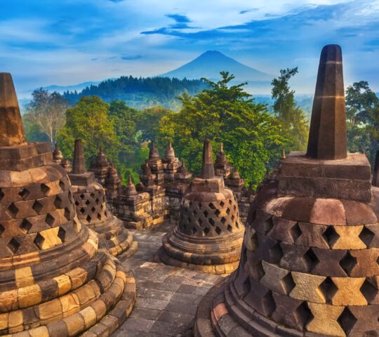 Ancient stone stupas at Borobudur Temple with a large volcanic peak and green forest in the distance.