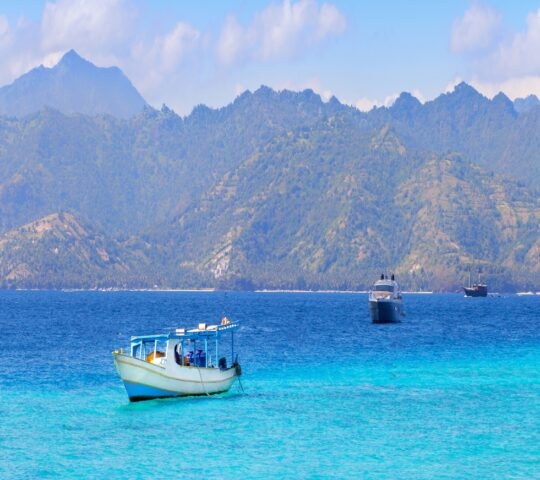 A white boat on clear blue water with several large, jagged mountain peaks rising in the background.