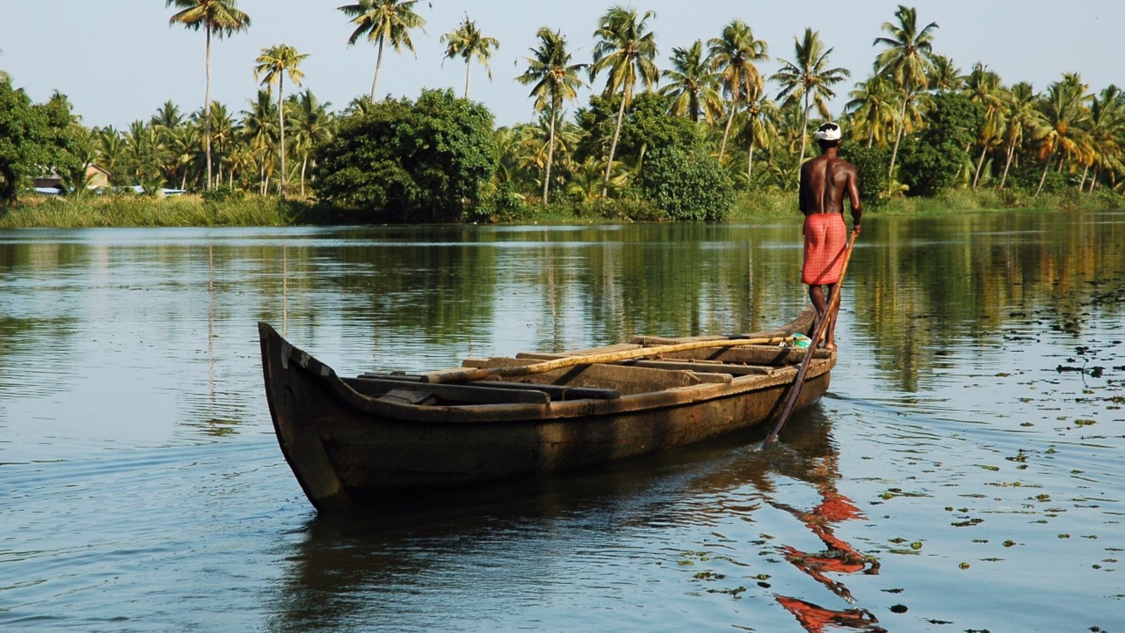 A man stands on the bow of a canoe with a long paddle steering it through palm-lined water in Kerala, India