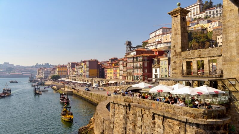 A sunny riverfront with outdoor cafe seating, historic tall buildings, and traditional wooden boats on the water.