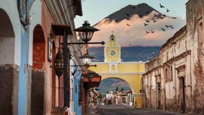 A yellow colonial arch over a cobblestone street with a large volcano and birds flying in the sky behind it.