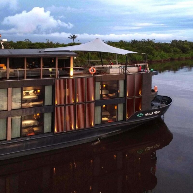 Side view of the Aqua Nera river cruise ship with lit windows reflecting on dark water near a lush forest shoreline.