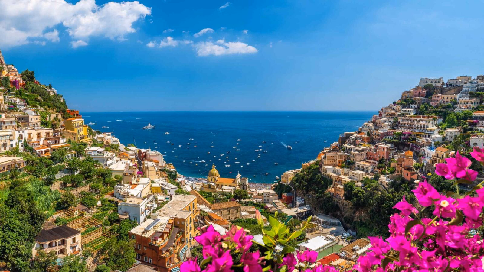 Colorful hillside buildings of Positano overlooking the Mediterranean Sea with pink bougainvillea in the foreground.