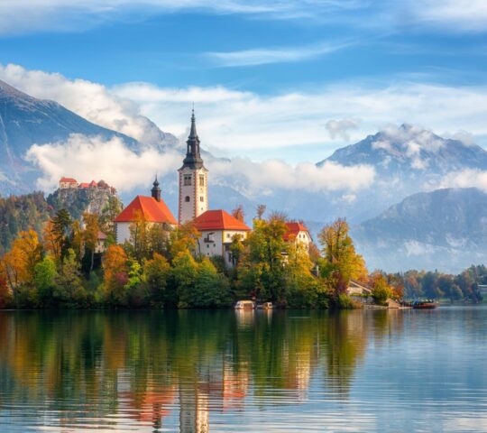 A white church with a tall steeple on a small island in the middle of a calm lake.