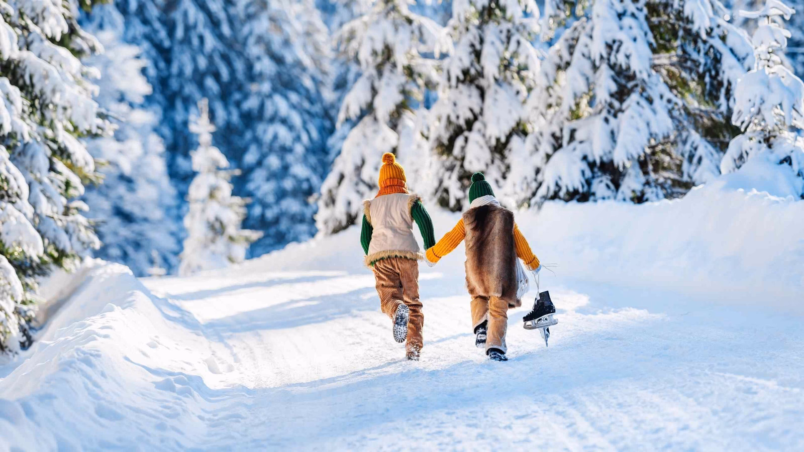 Two kids running away down a snowy forest road holding hands and carrying ice skates.