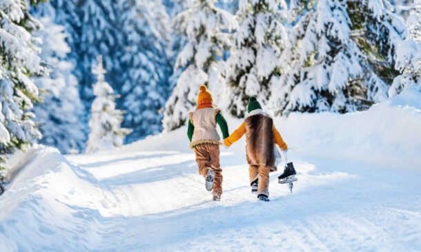 Two kids running away down a snowy forest road holding hands and carrying ice skates.