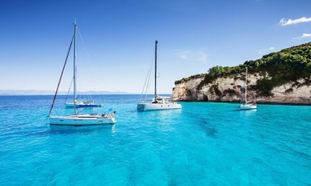 White sailboats floating on bright blue tropical water near a rocky coastline with green shrubbery.