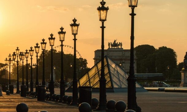 A row of silhouetted street lamps leads toward the Louvre pyramid and an archway at sunset.
