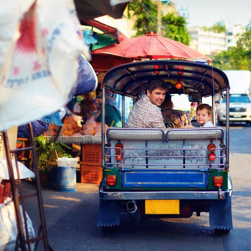 A man and a young boy looking back from the rear seat of a blue and green tuk-tuk on a city street.