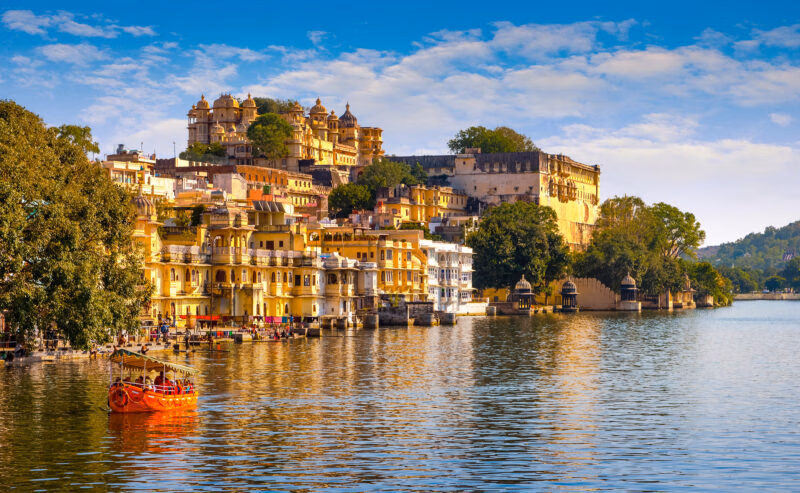 A large yellow palace complex sits on the edge of a lake with a small orange tour boat on the water.