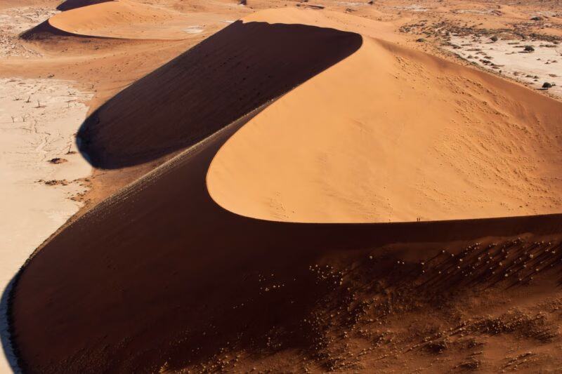 Aerial view of curved orange sand dunes with prominent dark shadows stretching across a vast desert floor.