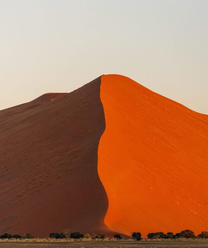 A tall sand dune with a sharp crest dividing bright orange sand from dark shadow against a light sky.
