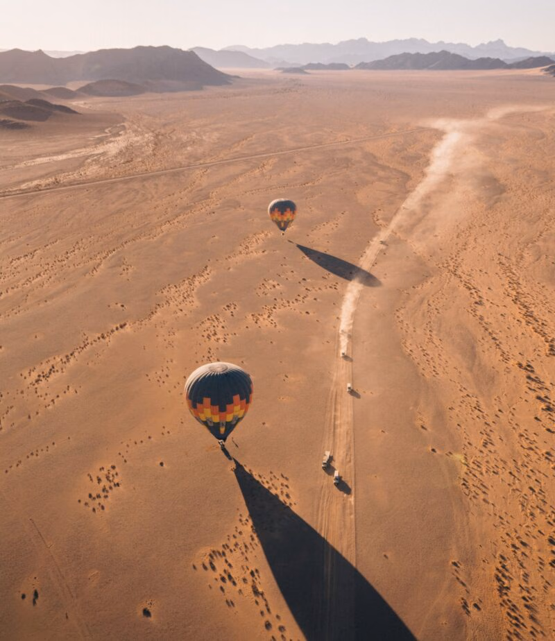 Aerial view of hot air balloons and vehicles on a dirt track in a vast desert landscape.