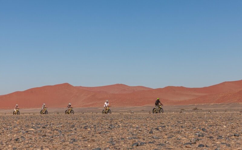 Five people riding bicycles across a rocky desert floor with red sand dunes under a blue sky.