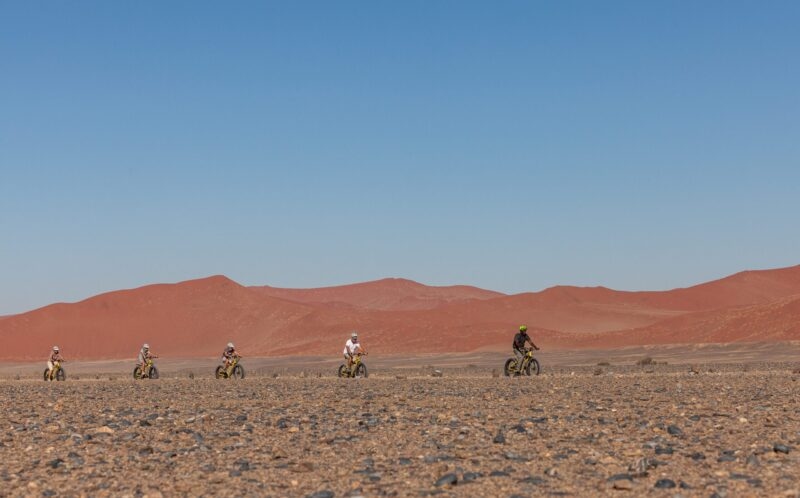 Five people riding bicycles across a rocky desert floor with red sand dunes under a blue sky.