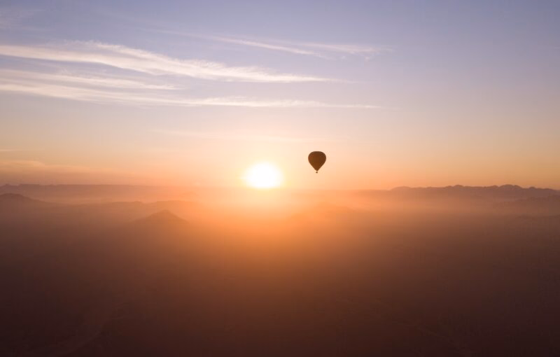 Silhouette of a hot air balloon in a bright orange sunrise sky over misty mountains.