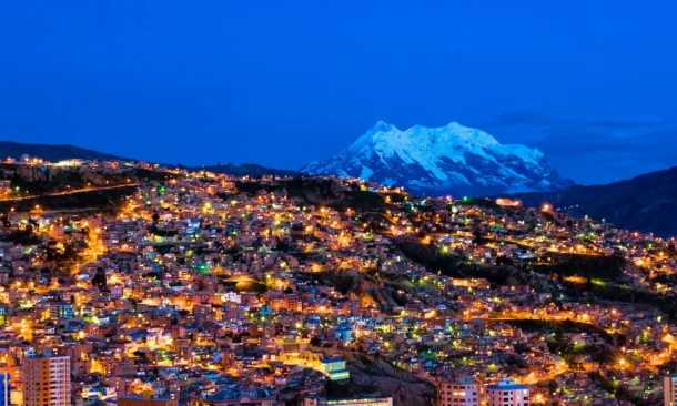 Warm city lights across a steep hillside leading up to a massive snow-capped mountain under a dark blue twilight sky.