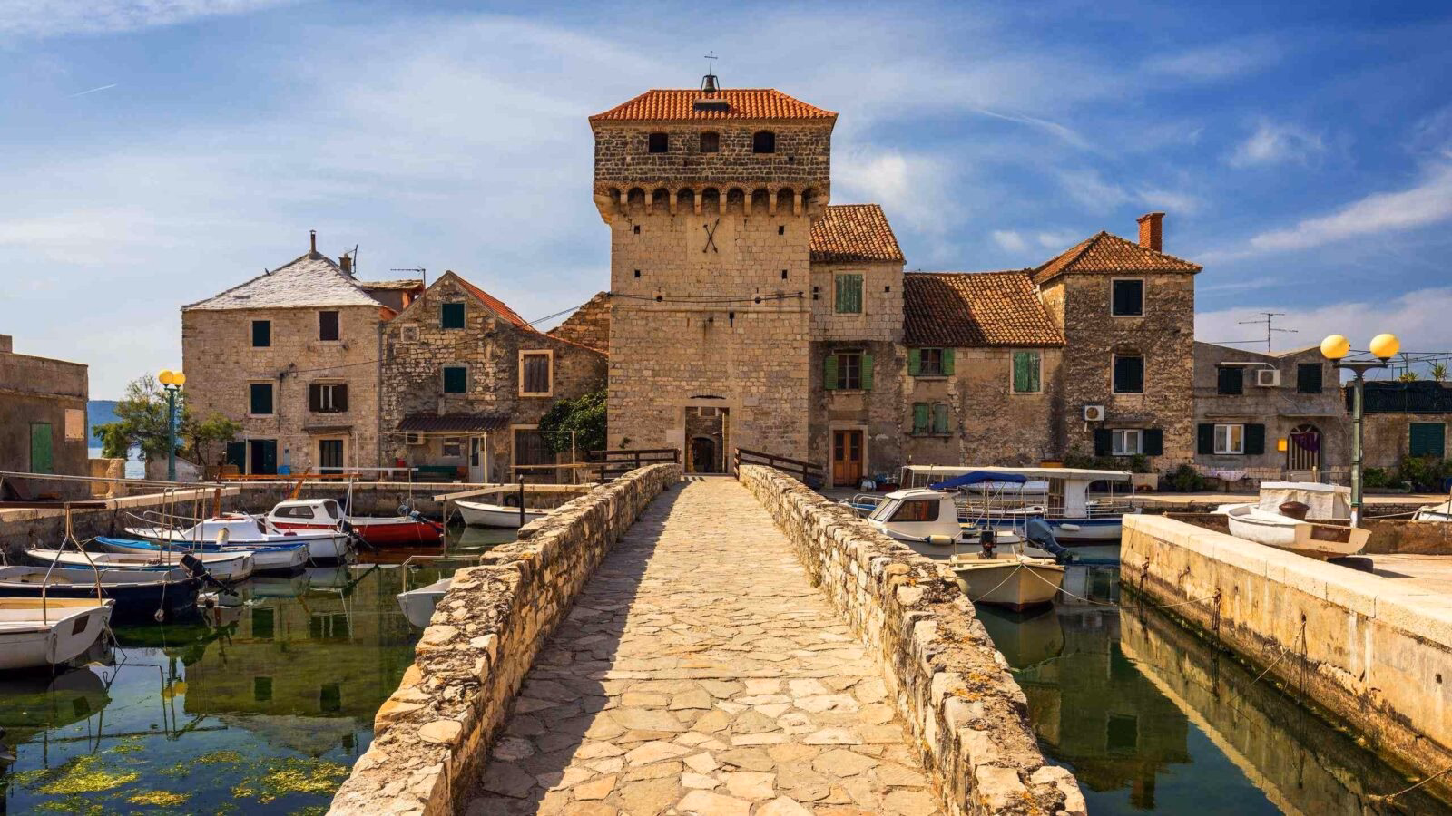 A stone bridge leading to a medieval tower in a harbor filled with small white boats and historic buildings.