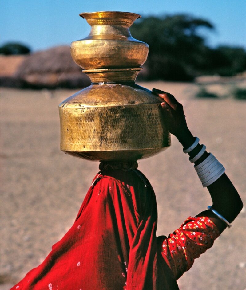 A woman in a red head scarf carrying a large gold pot on her head, holding it with one hand, on which she wears many white bangles