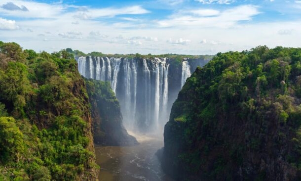 High angle view of a wide waterfall flowing between two tall, tree-covered cliffs into a river below.